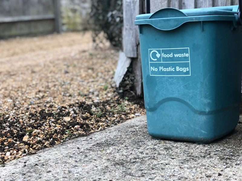 A compost bin in front of a wooden fence. The bin says Food Waste, No Plastic Bags