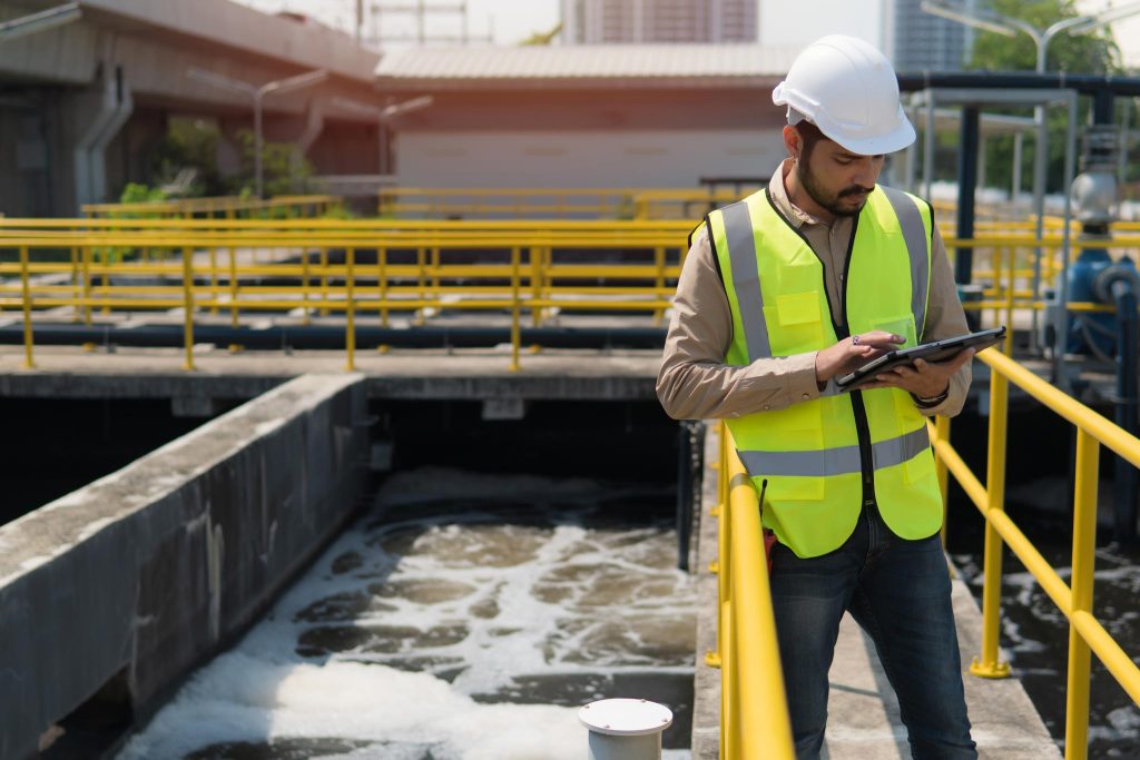 Service engineer checking on waste water treatment plant with pump on background. Engineer is using an iPad.